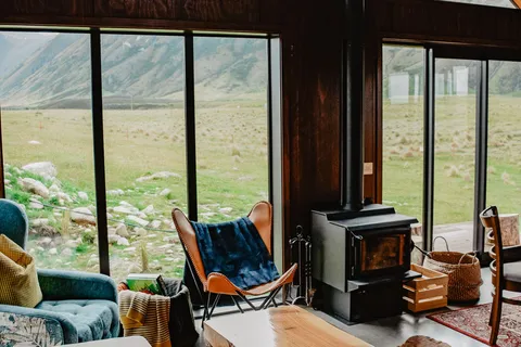 Wood-panelled living room with a wood stove, armchairs and tall windows facing a green valley