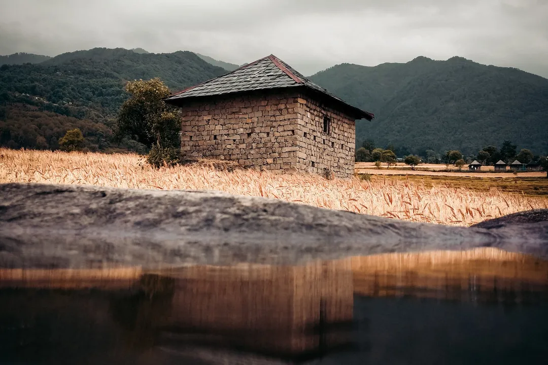 Stone house with a slate roof beside a golden field and still water, with forested hills behind