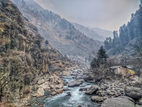 Clear mountain river flowing over boulders through a steep wooded valley with a small house
